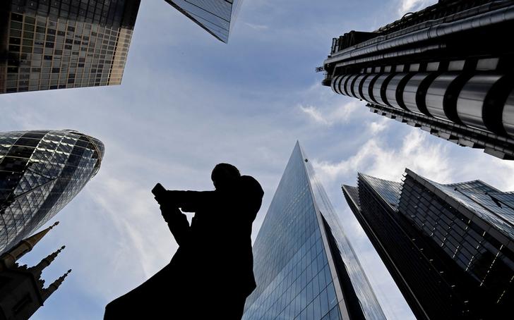 A worker looks at their phone as they walk past The Gherkin, Lloyds, and other office buildings in the City of London